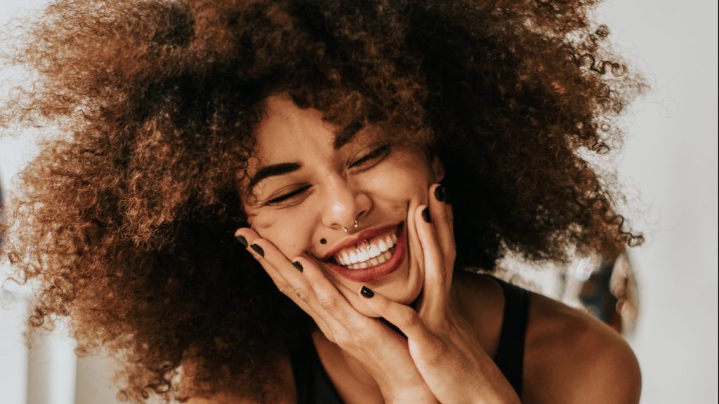 Young Adult Woman With Afro And Nose Piercing Smiling With Hands Pressed To Her Face And Sitting In Chair