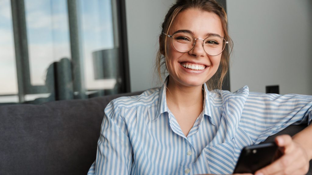 Young woman in glasses and striped shirt smiling with phone cosmetic dentist Portage MI